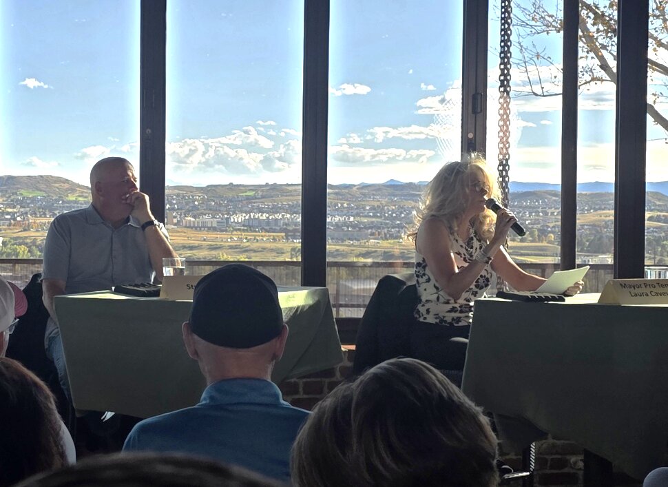 Mayor Pro Tem Laura Cavey speaking into a microphone at the candidate panel table during the Castle Rock Chamber forum, with a packed audience in the foreground and a panoramic view of Castle Rock through the windows behind her