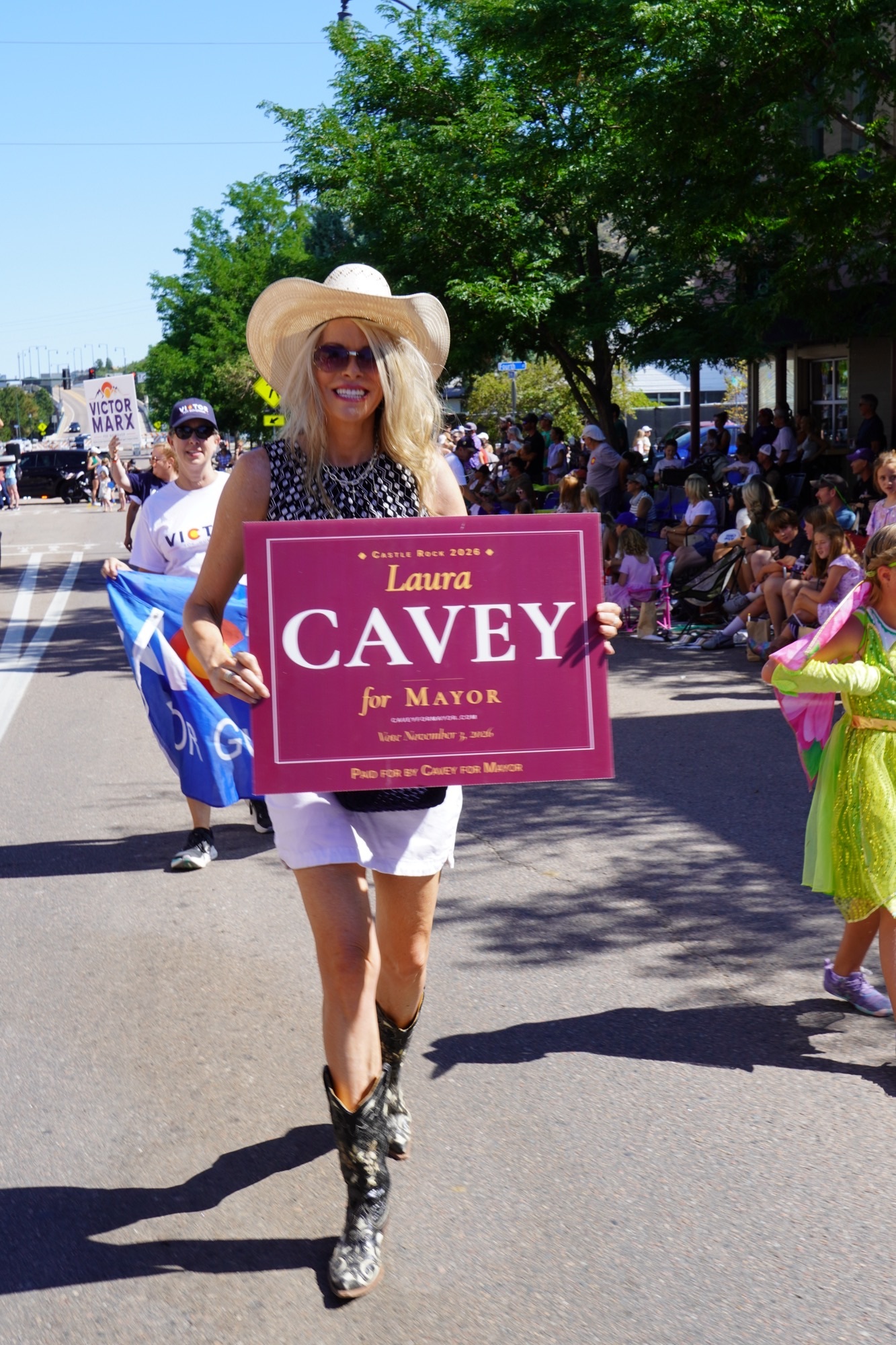 Laura Cavey walking the Douglas County Fair Parade route through downtown Castle Rock in a cowboy hat and boots, carrying her Laura Cavey for Mayor sign past crowds lining the street