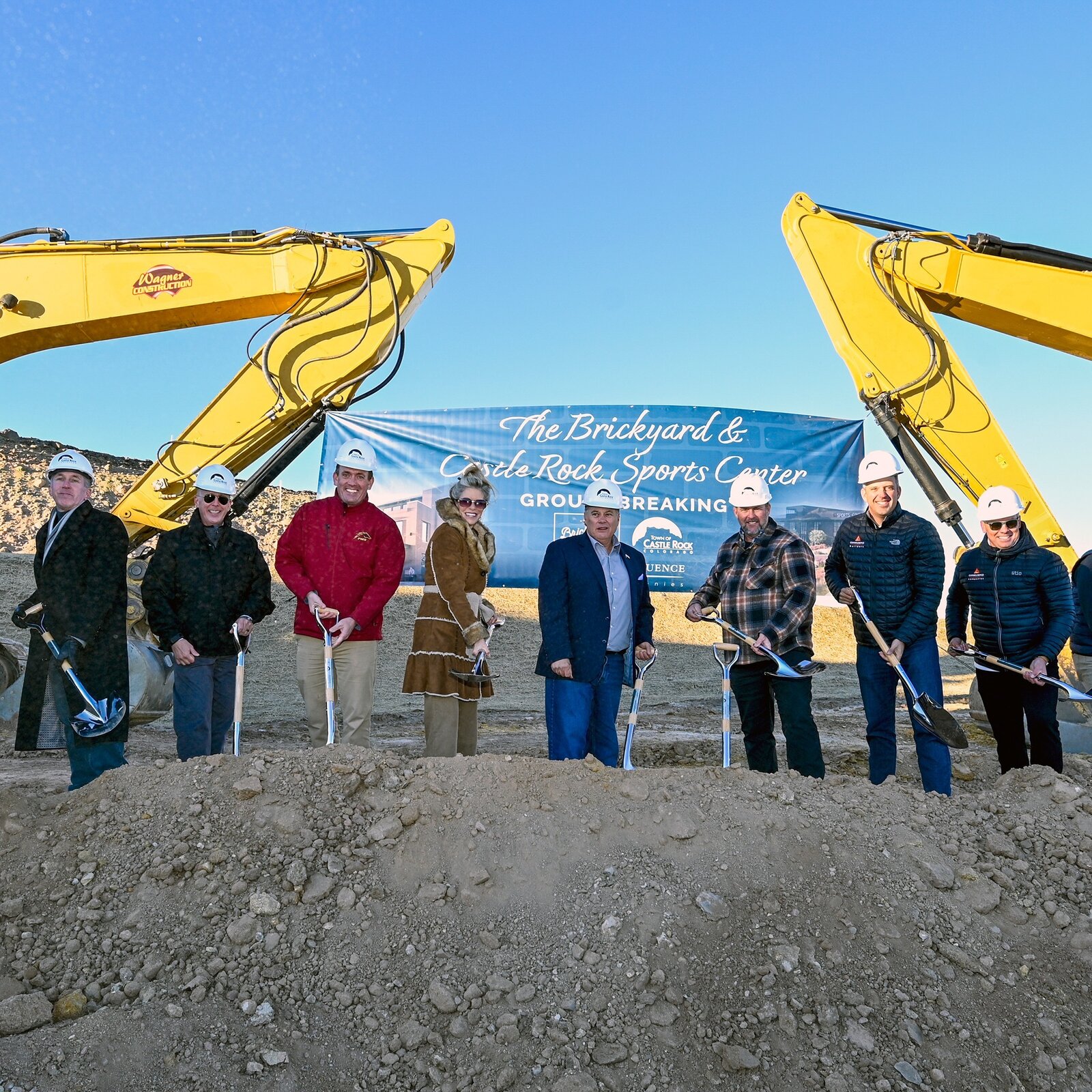 Laura Cavey with Town leaders and project partners in hard hats, turning dirt with silver shovels between two excavators at the Brickyard and Castle Rock Sports Center groundbreaking